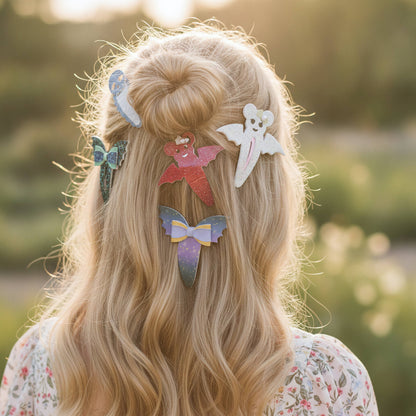 A person viewed from behind with long blonde hair styled in a half-up bun, wearing several decorative hair clips shaped like a ghost, butterflies, and abstract figures against a softly blurred outdoor background.