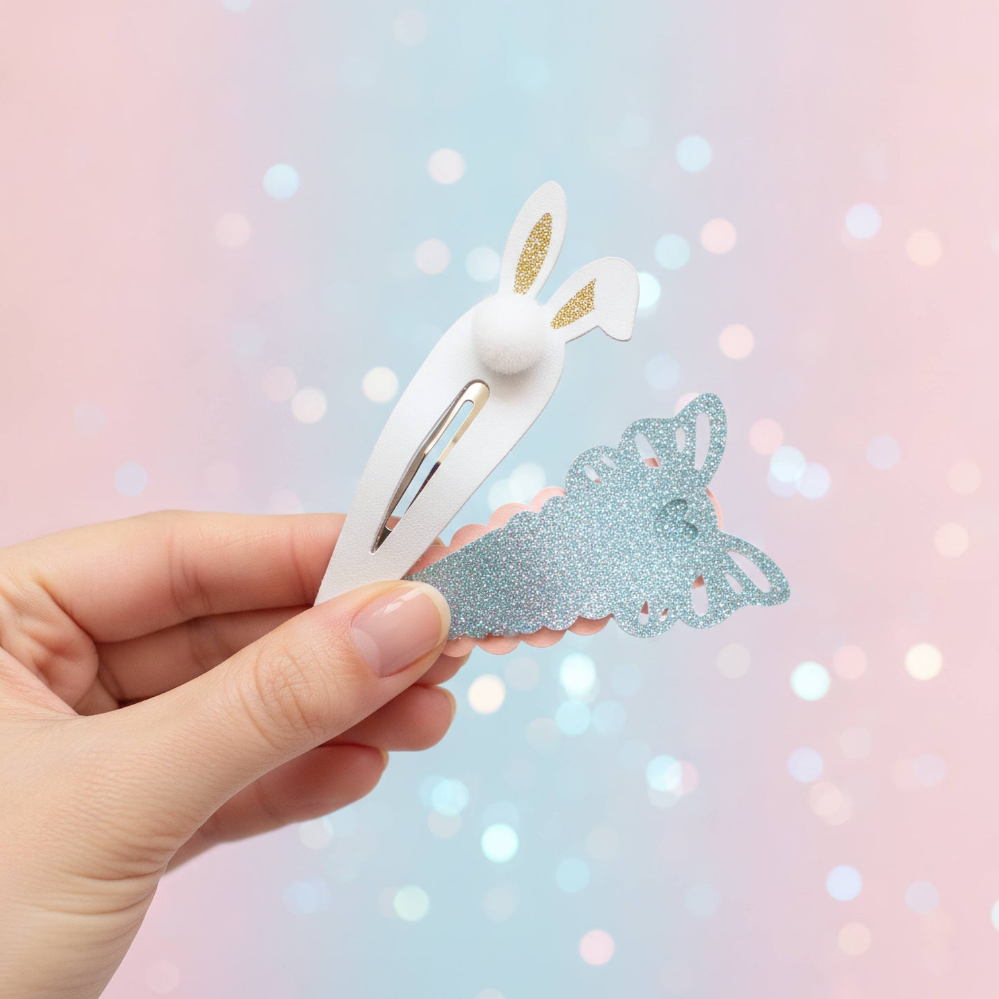 A hand holds a white plastic hair clip shaped like bunny ears next to a glittery light blue butterfly-shaped hair tie against a pastel pink and blue bokeh background.