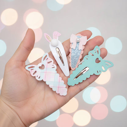 Four pastel hair clips held in a hand against a soft-focus bokeh background. The clips include a white bunny clip with pink glitter ears, a pink glitter bow clip with a pearl, a mint green butterfly clip, and a white plaid ribbon clip.