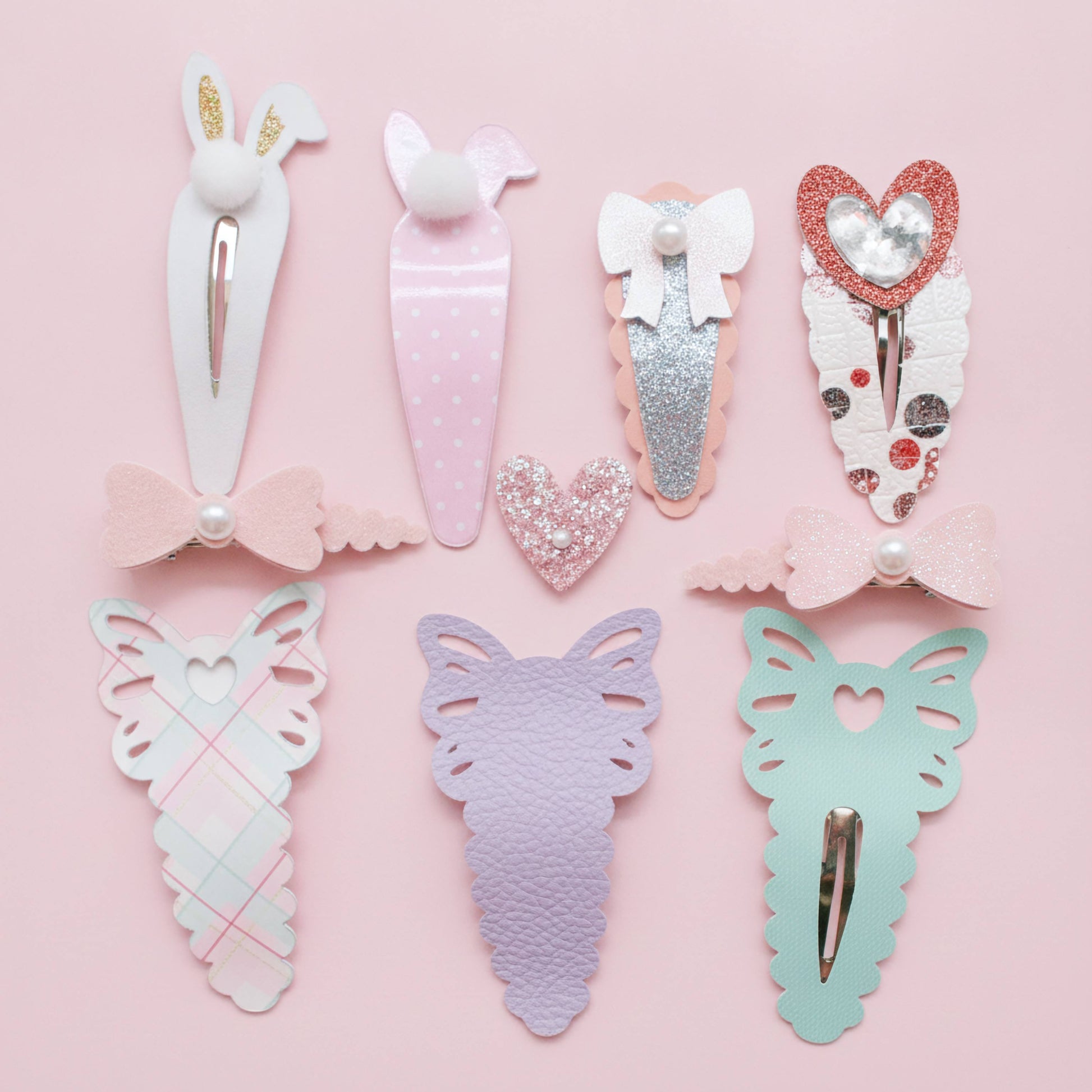 A set of eight pastel hair clips arranged on a light pink background. The clips feature bunny, carrot, bow, heart, and butterfly designs in soft pastel colors including pink, lavender, mint green, white, and glittery rose gold.