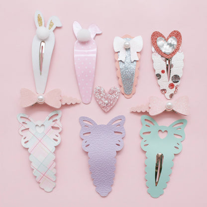 A set of eight pastel hair clips arranged on a light pink background. The clips feature bunny, carrot, bow, heart, and butterfly designs in soft pastel colors including pink, lavender, mint green, white, and glittery rose gold.