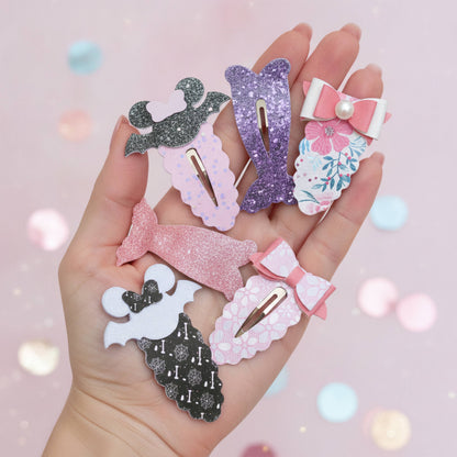 A hand holding six mermaid-themed hair clips in various glittery and patterned designs, set against a soft pink background with bokeh light effects.