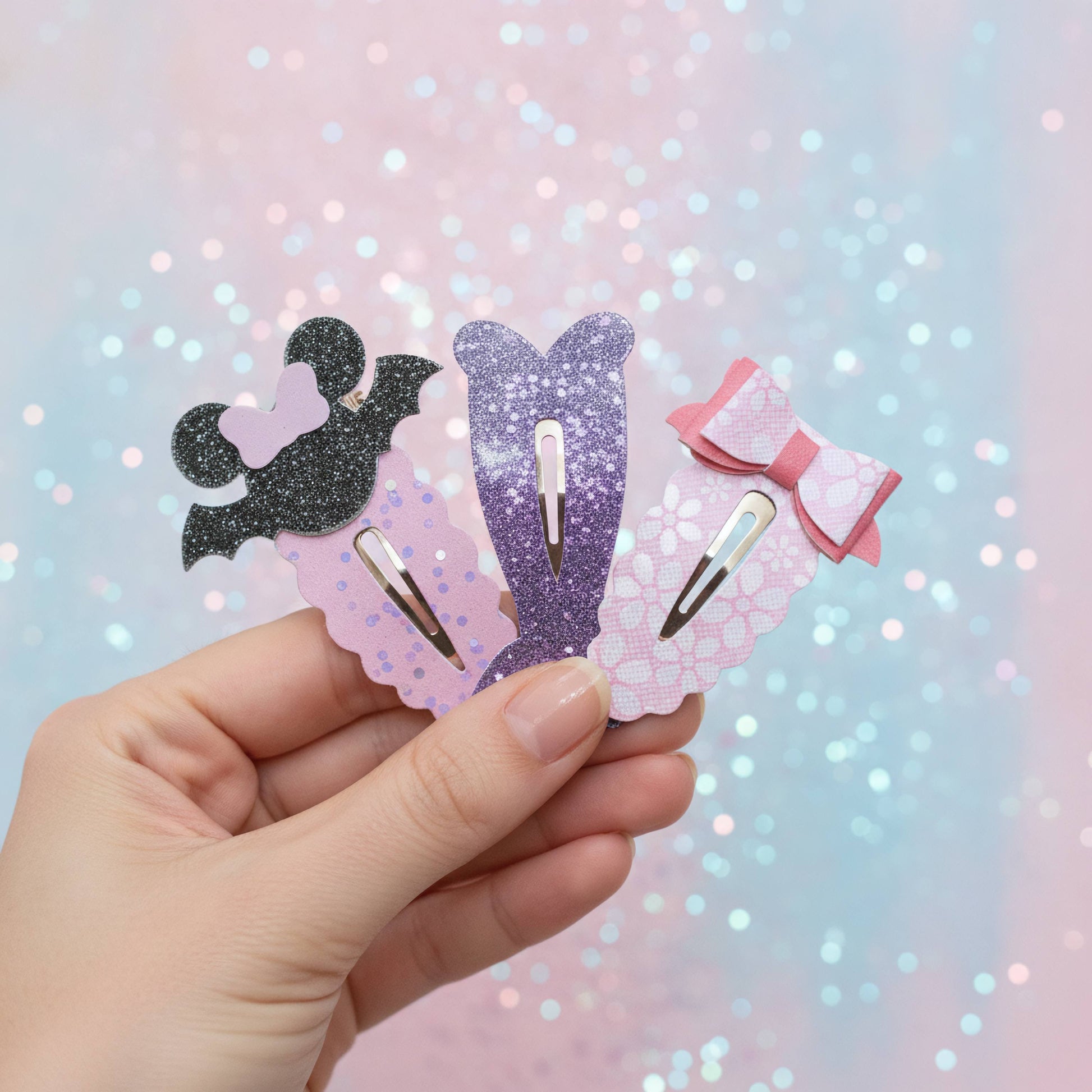 A hand holds three cute mermaid-themed hair clips against a pastel pink and blue glittery background. The clips feature a black bat silhouette, a purple mermaid tail, and a pink bow on scalloped pastel backgrounds.