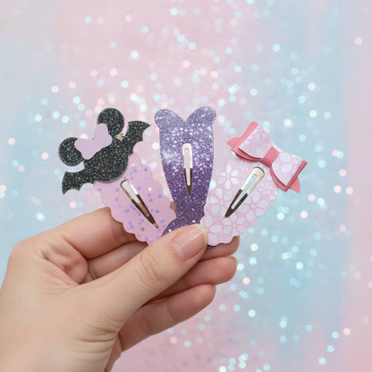 A hand holds three cute mermaid-themed hair clips against a pastel pink and blue glittery background. The clips feature a black bat silhouette, a purple mermaid tail, and a pink bow on scalloped pastel backgrounds.