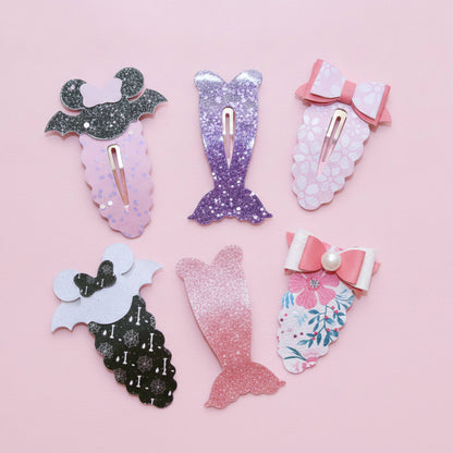 Six mermaid-themed hair clips arranged on a solid pink background. The clips feature various designs including glittery mermaid tails, floral mermaid tails, and mermaid tails with bows, in colors like pink, purple, black, and white.