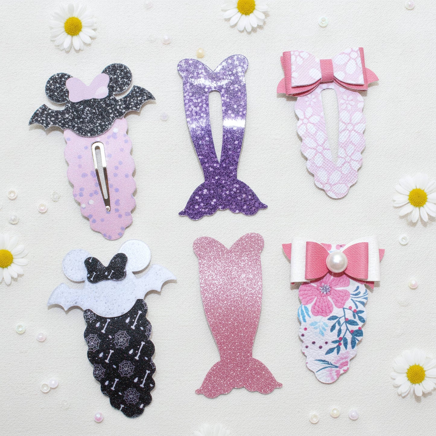 Six mermaid-themed hair clips arranged on a white background with small daisy props. The clips include glittery and patterned designs in pink, purple, black, and white, with bows and mermaid tail motifs.