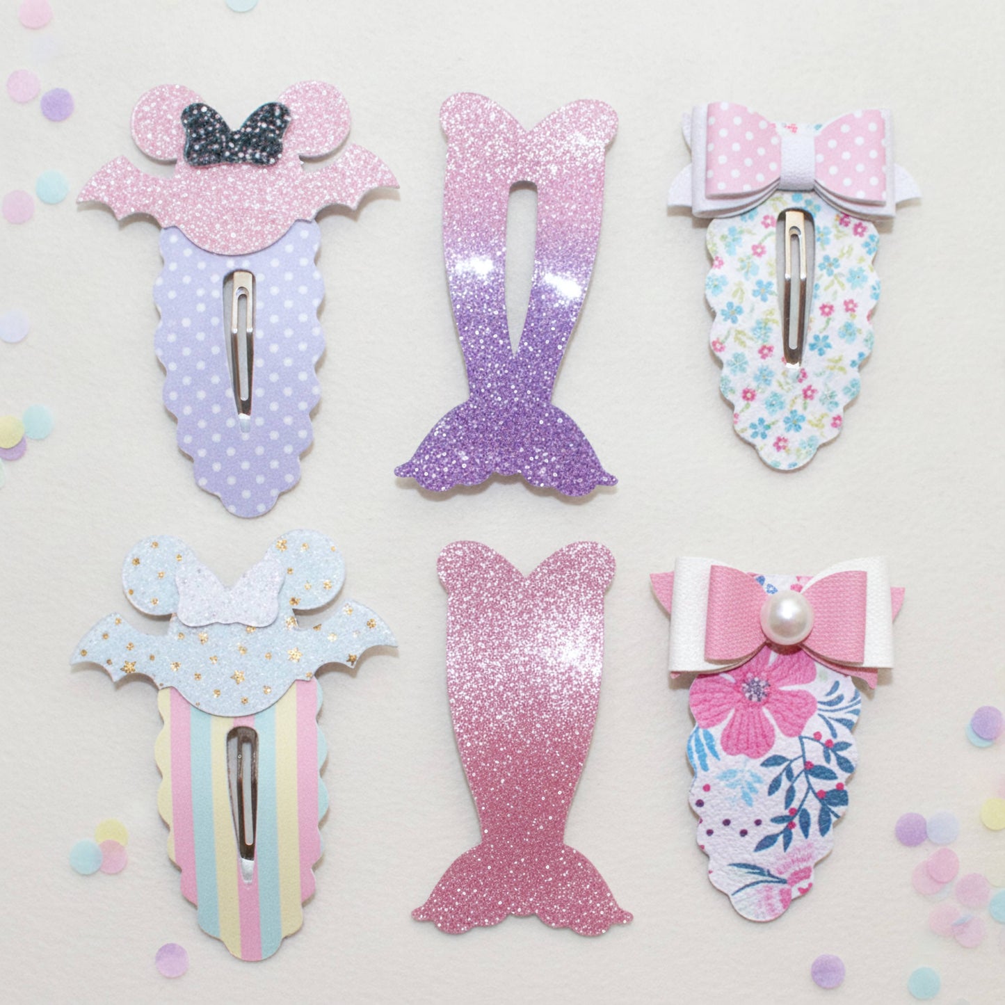 Six mermaid-themed hair clips arranged on a light cream background with scattered pastel confetti. The clips feature glittery pink and purple mermaid tails, floral prints, pastel stripes, bows, and polka dots in pink, blue, white, and yellow.