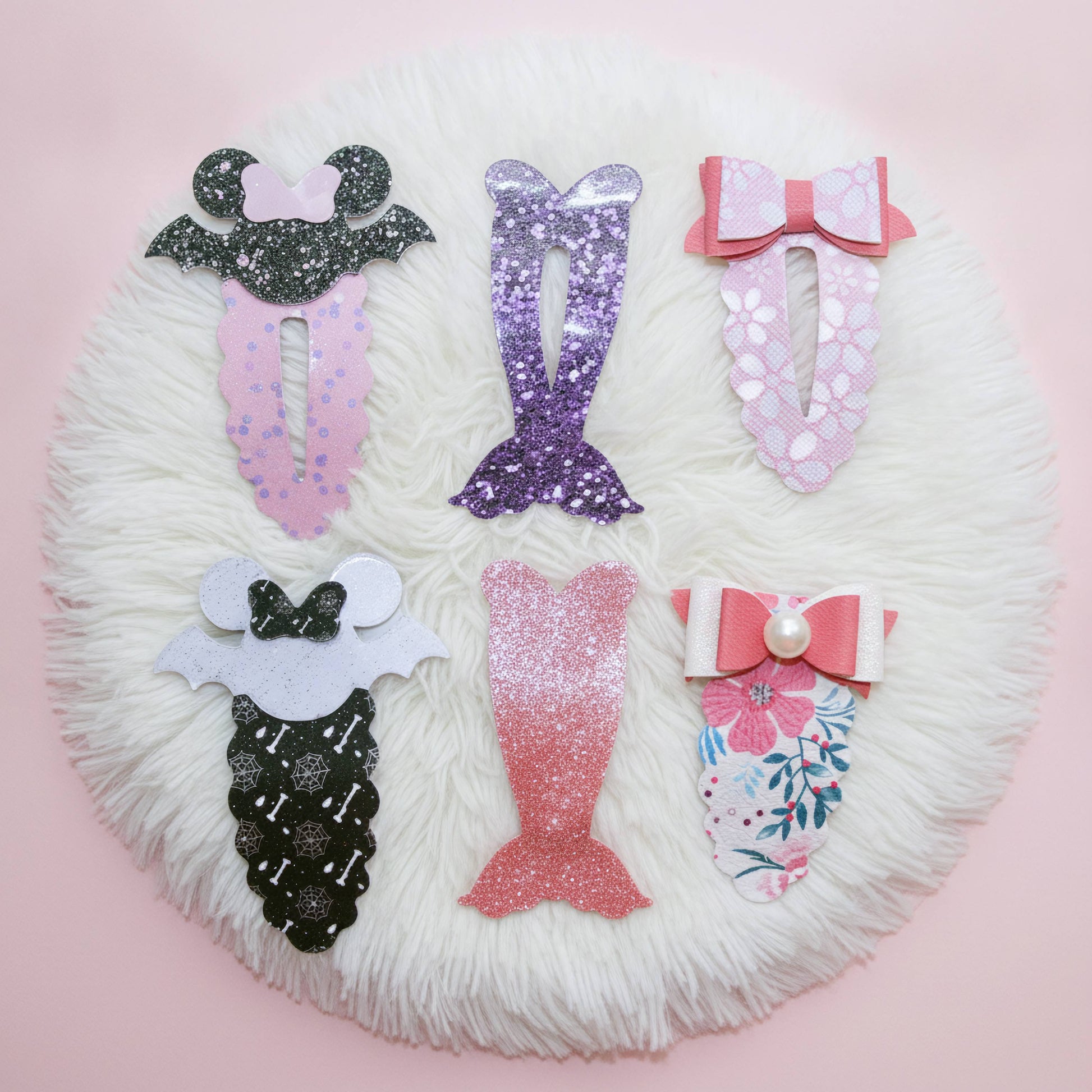 Six mermaid-themed hair clips arranged on a round white faux-fur rug against a light pink background. The clips feature glittery, glitter-finish, floral, and patterned designs in pink, purple, black, white, and floral colors.