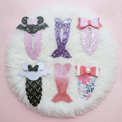 Six mermaid-themed hair clips arranged on a round white faux-fur rug against a light pink background. The clips feature glittery, glitter-finish, floral, and patterned designs in pink, purple, black, white, and floral colors.