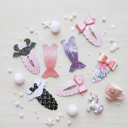 A flat-lay collection of mermaid-themed hair clips and hair bows in various colors and patterns, including pink, purple, black, and floral designs, arranged on a light wood background with decorative stars, pearls, and a small teacup.