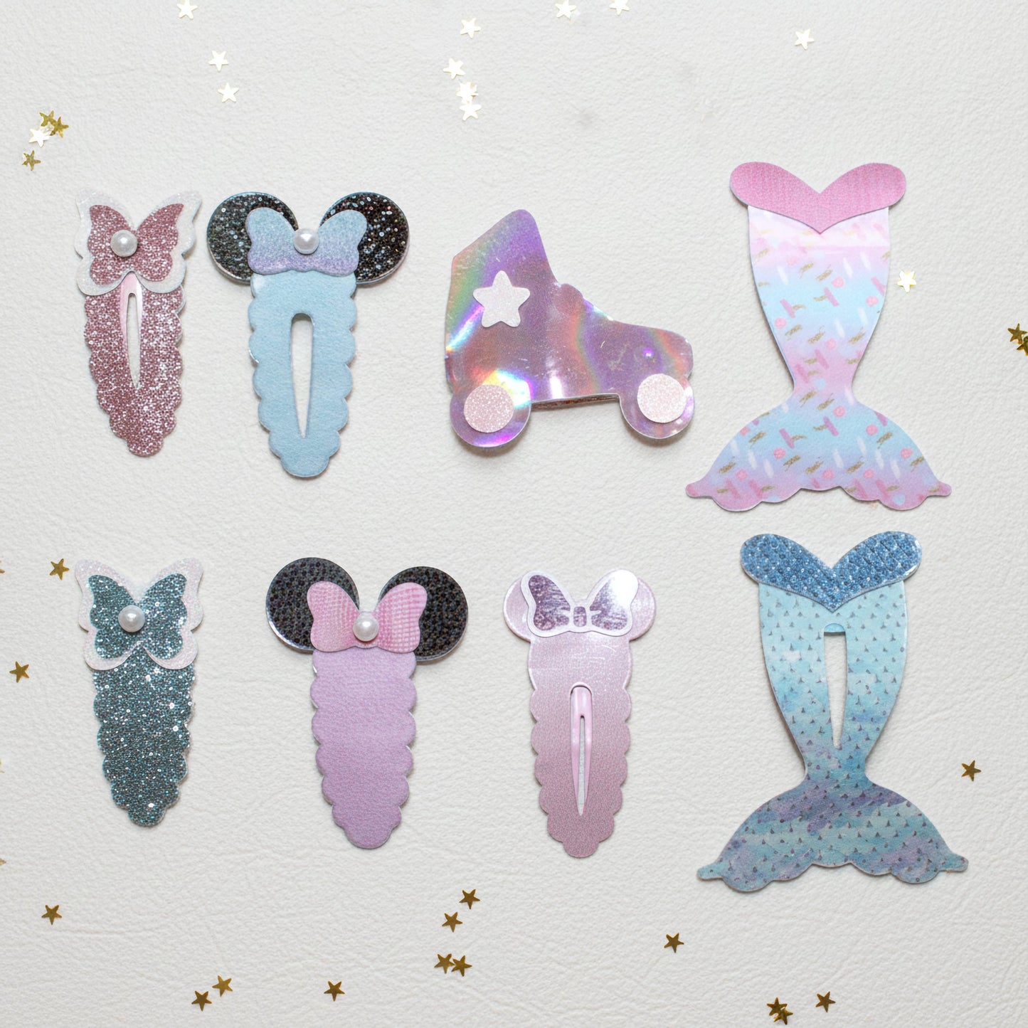 Six mermaid-themed hair clips arranged on a textured white background with scattered gold star confetti. The clips include glittery pink, glittery blue, holographic roller skate, and mermaid tail designs in pastel pink, lavender, and blue tones.
