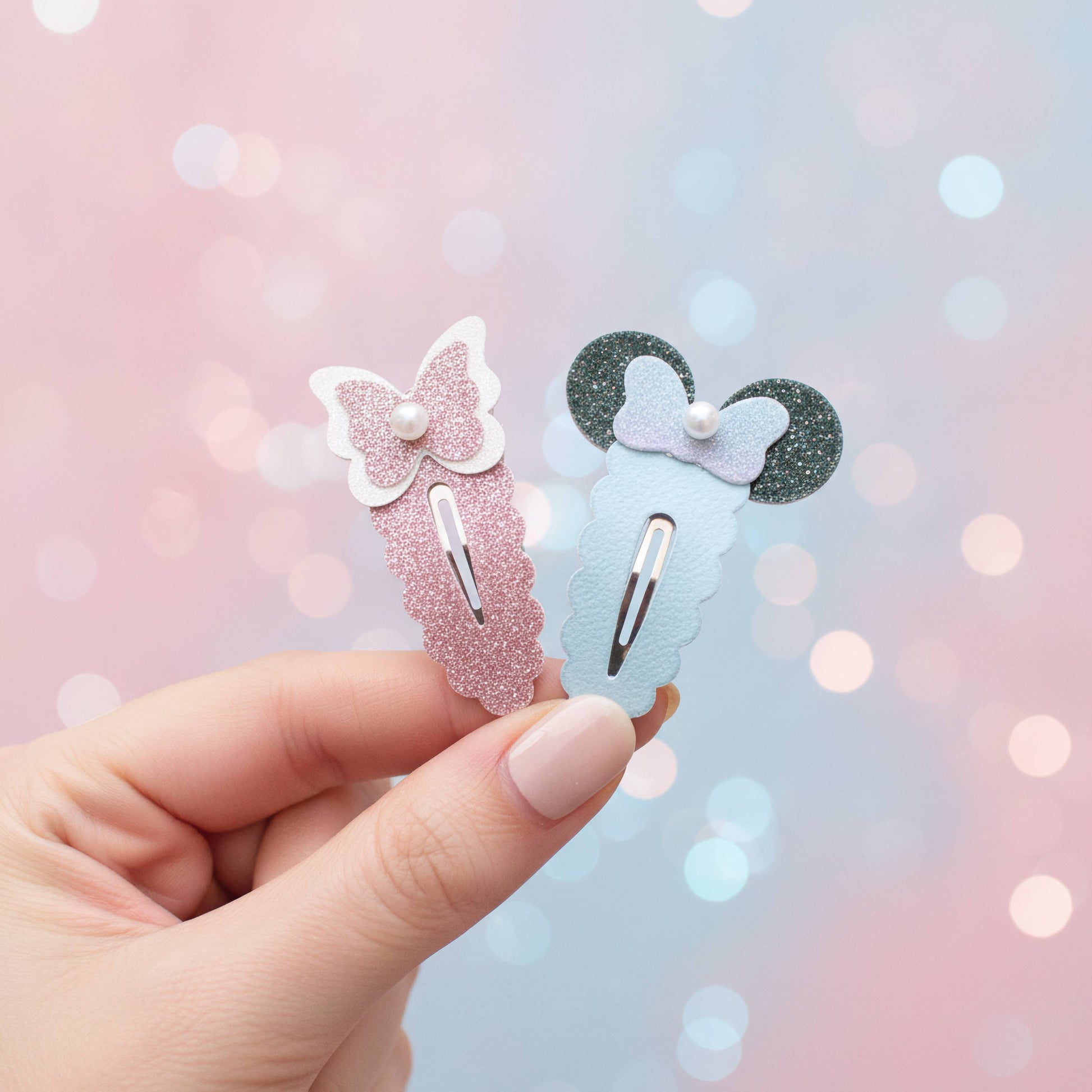 A hand holds two decorative hair clips against a soft pink and blue bokeh background. One clip is light pink glitter with a white bow and pearl, and the other is light blue glitter with a lavender bow and dark glitter ears.