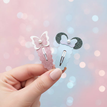 A hand holds two decorative hair clips against a soft pink and blue bokeh background. One clip is light pink glitter with a white bow and pearl, and the other is light blue glitter with a lavender bow and dark glitter ears.