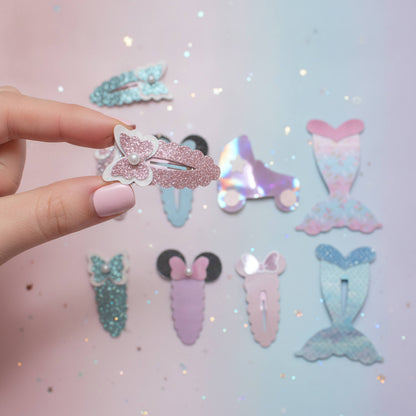 A hand with pale pink nails holds a pink glitter mermaid tail hair clip against a pastel pink and blue glittery background, with several other mermaid-themed hair clips arranged behind it.