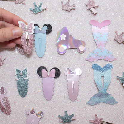 A collection of pastel mermaid and fairy-themed hair clips arranged on a sparkly pink background, with a hand holding one clip on the left.