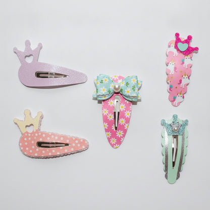 Five decorative hair clips arranged on a plain light background. The set includes a lavender glitter crown clip, a pink carrot clip with white polka dots and a small cream crown, a mint bow clip with a pink daisy-patterned body, a pink unicorn clip with a crown topper, and a pale mint glitter clip with a crystal-like accent.