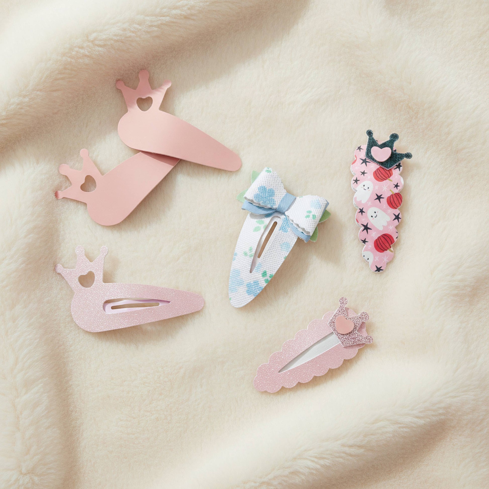 Five decorative hair clips arranged on a soft cream fabric background. The set includes pastel pink clips with crown motifs, a white clip with blue floral print and a light blue bow, a pink strawberry-themed clip, and a pink glitter clip with a small crown accent.