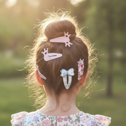 A child viewed from behind with brown hair in a high bun, styled with several pastel hair clips including a pink bunny clip, a white horse clip, a blue bow clip, and a floral hair accessory, set against a warm, sunlit outdoor background.