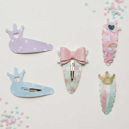 A set of five pastel unicorn and princess-themed hair clips arranged on a light off-white background with scattered star confetti. The clips feature pastel purple, blue, pink, and mint green colors, floral patterns, glittery pink bows, and gold crown accents.