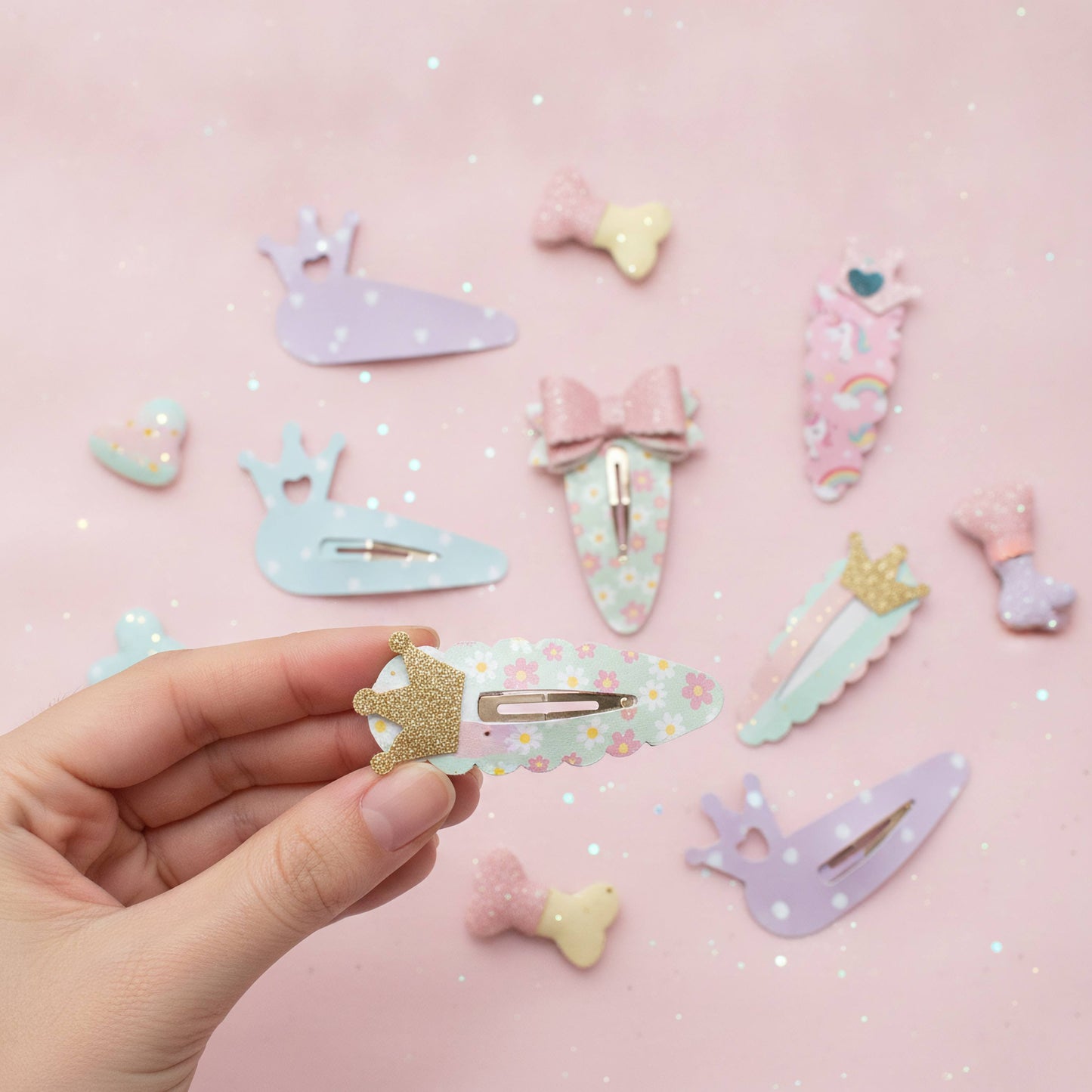 A hand holds a pastel floral hair clip with a gold glitter crown, surrounded by other pastel-colored hair clips shaped like bows, whales, and unicorns on a pink glittery background.