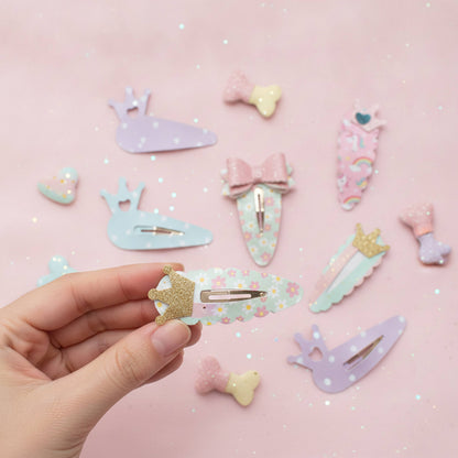 A hand holds a pastel floral hair clip with a gold glitter crown, surrounded by other pastel-colored hair clips shaped like bows, whales, and unicorns on a pink glittery background.