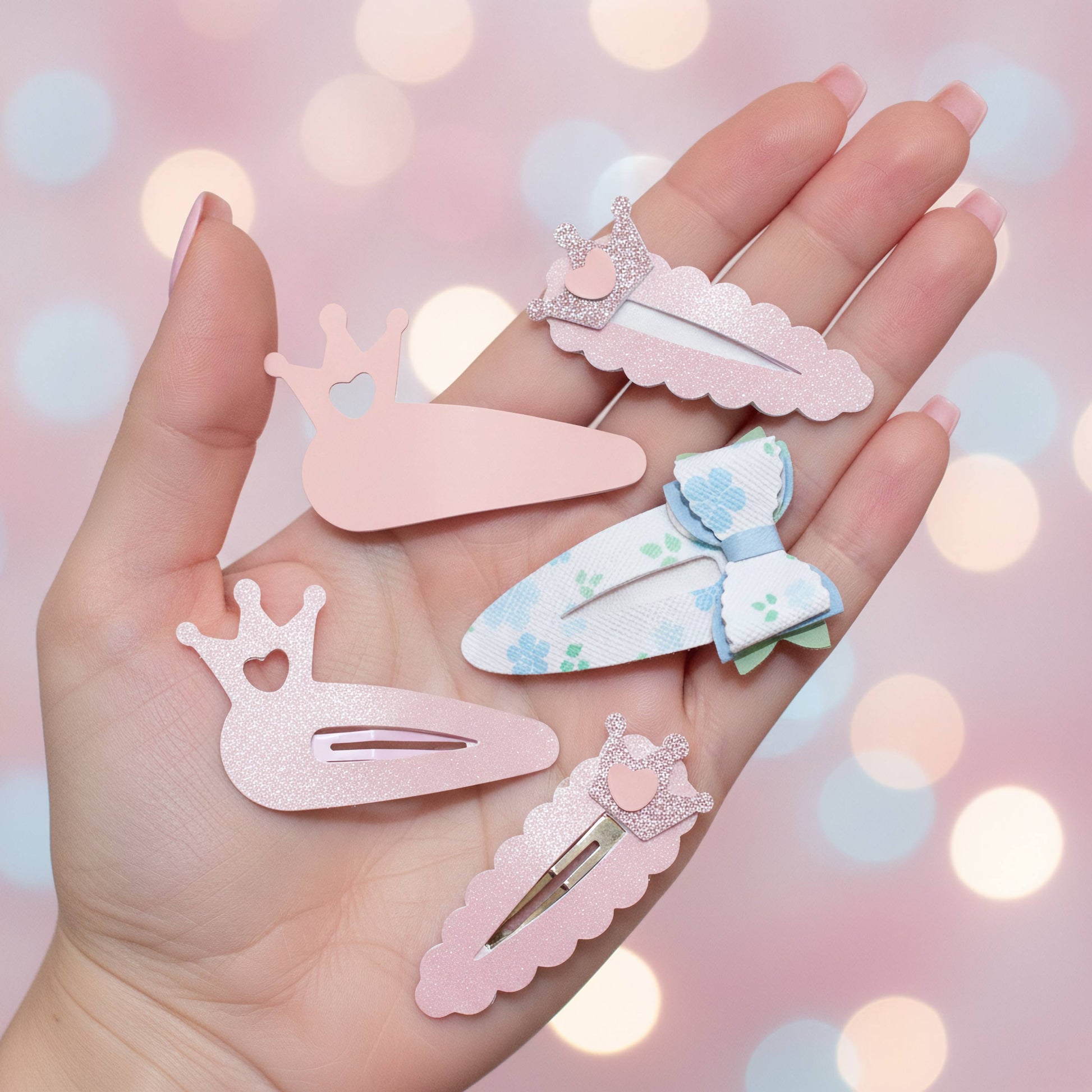 Four pastel hair clips held in a hand against a soft pink bokeh background. The clips include a glittery pink crown clip, a light pink scalloped clip with a small heart, a white clip with a blue bow and green floral pattern, and a pink scalloped clip with a glittery crown and heart.
