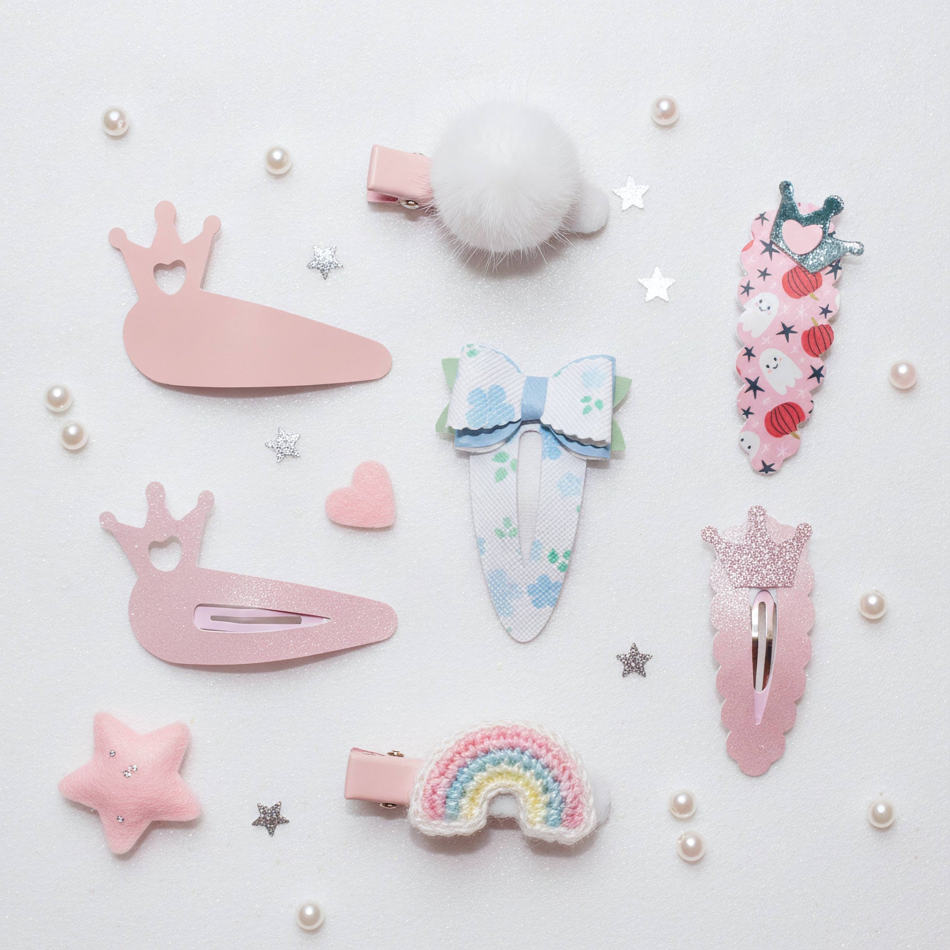 A set of pastel pink and multicolor hair clips arranged on a white background with decorative pearls and star stickers. The clips include a fluffy pom-pom clip, two pink bunny clips, a floral bow clip, a strawberry-themed clip, a glittery feather clip, a rainbow clip, and a small star clip.