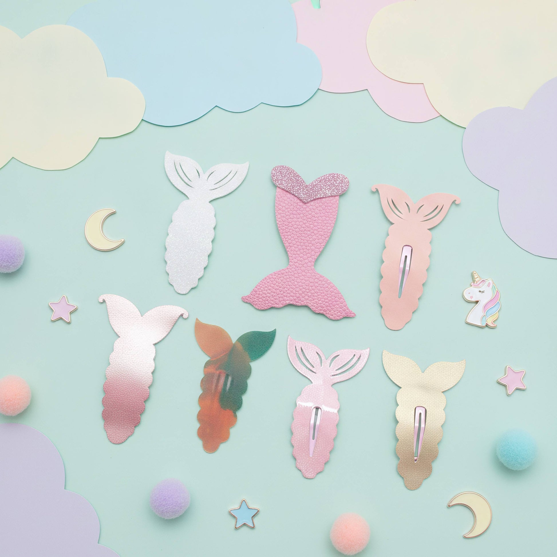A collection of pastel mermaid tail hair clips arranged on a light blue background with decorative paper clouds, stars, moons, and pom-poms. The clips come in various colors including pink, white, gold, and iridescent gradient finishes.