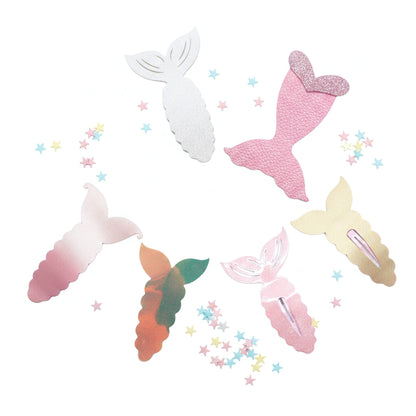Six mermaid tail hair clips in various designs including textured pink, glittery gold, ombre pink, gradient green-orange, and solid white, arranged on a white background with scattered pastel star confetti.