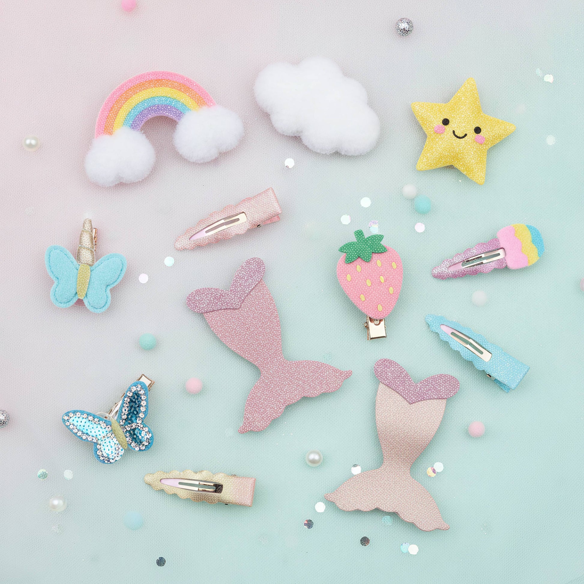A flat-lay collection of pastel hair clips and hair accessories on a light mint blue background with scattered glitter and beads. The set includes a rainbow with clouds, fluffy white clouds, a yellow glitter star, a blue glitter butterfly, a pink glitter mermaid tail, a strawberry clip, a pink and blue glitter ice cream cone clip, and several pastel-colored hair clips.