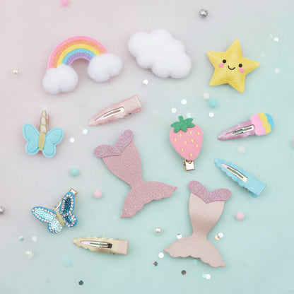 A flat-lay collection of pastel hair clips and hair accessories on a light mint blue background with scattered glitter and beads. The set includes a rainbow with clouds, fluffy white clouds, a yellow glitter star, a blue glitter butterfly, a pink glitter mermaid tail, a strawberry clip, a pink and blue glitter ice cream cone clip, and several pastel-colored hair clips.