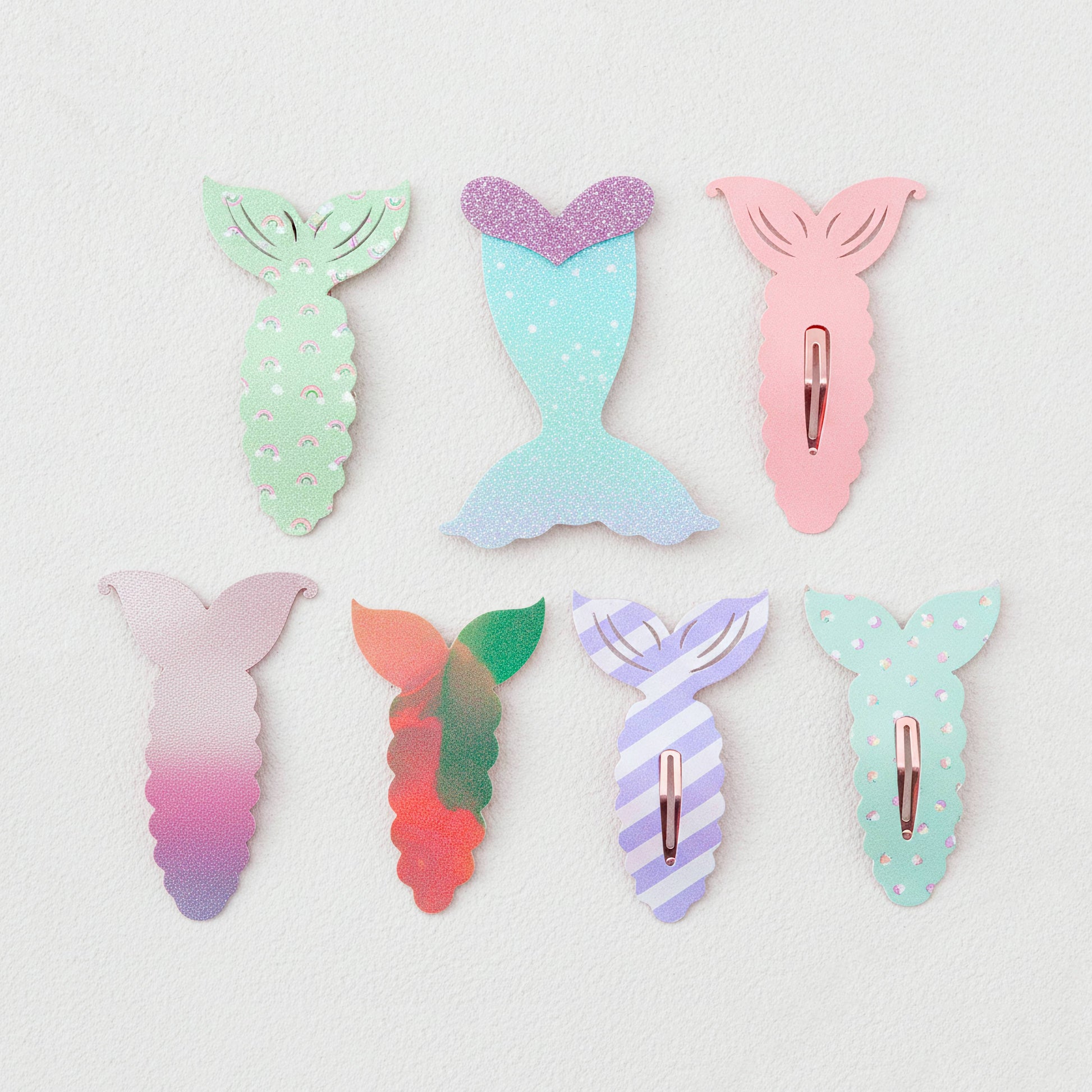 Six mermaid tail hair clips in pastel colors, arranged in two rows on a textured off-white background. The clips feature various designs including glitter, stripes, polka dots, and gradients.