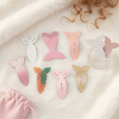 A set of pastel mermaid tail hair clips arranged on a soft white faux-fur surface, with a small pink drawstring pouch and a jar of clips nearby. The clips come in white, pink, peach, green-orange, and gold tones.