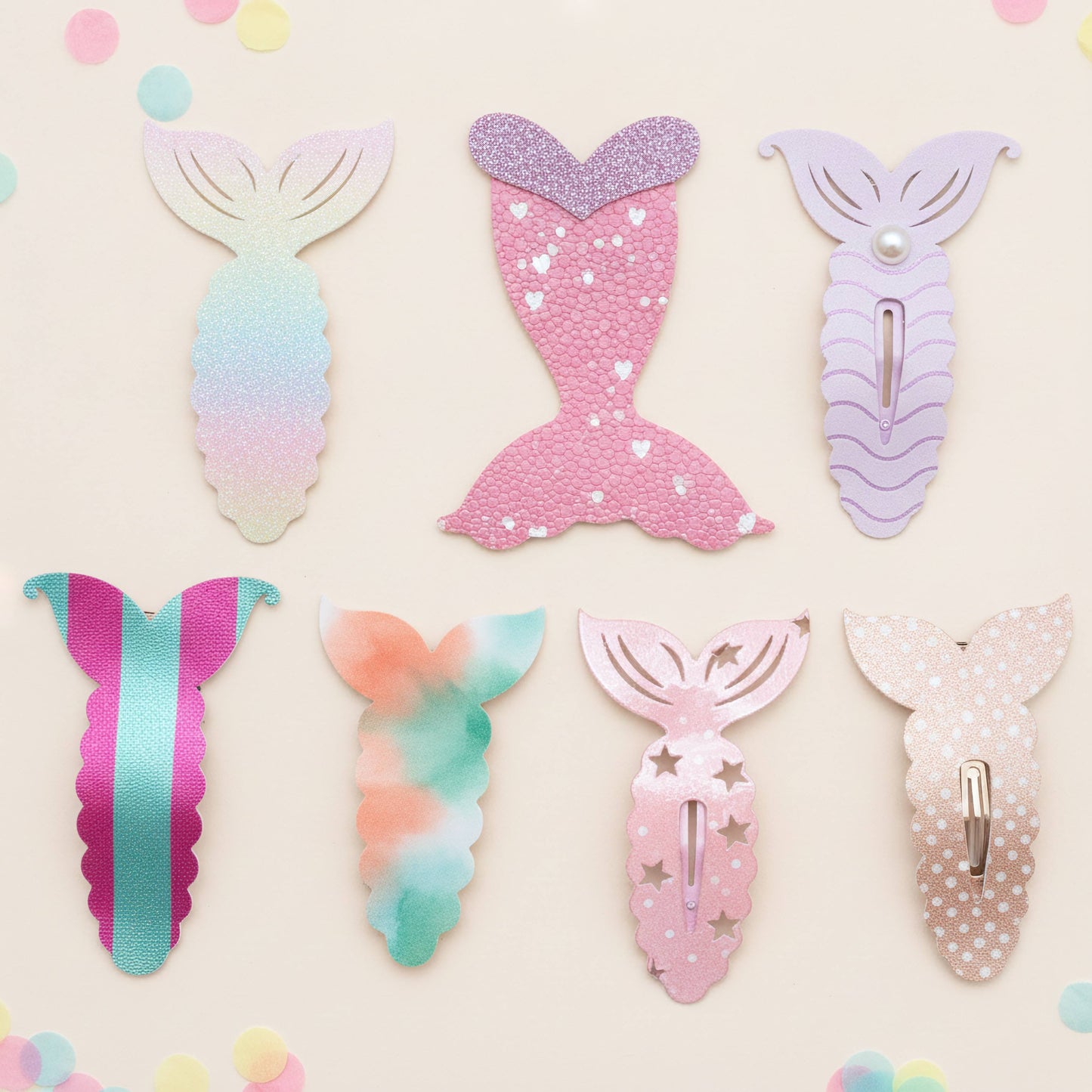 Six mermaid tail hair clips in pastel colors arranged on a light cream background with scattered confetti. The clips include gradient, glitter, striped, tie-dye, star, and polka-dot designs in pink, purple, mint, peach, and white.