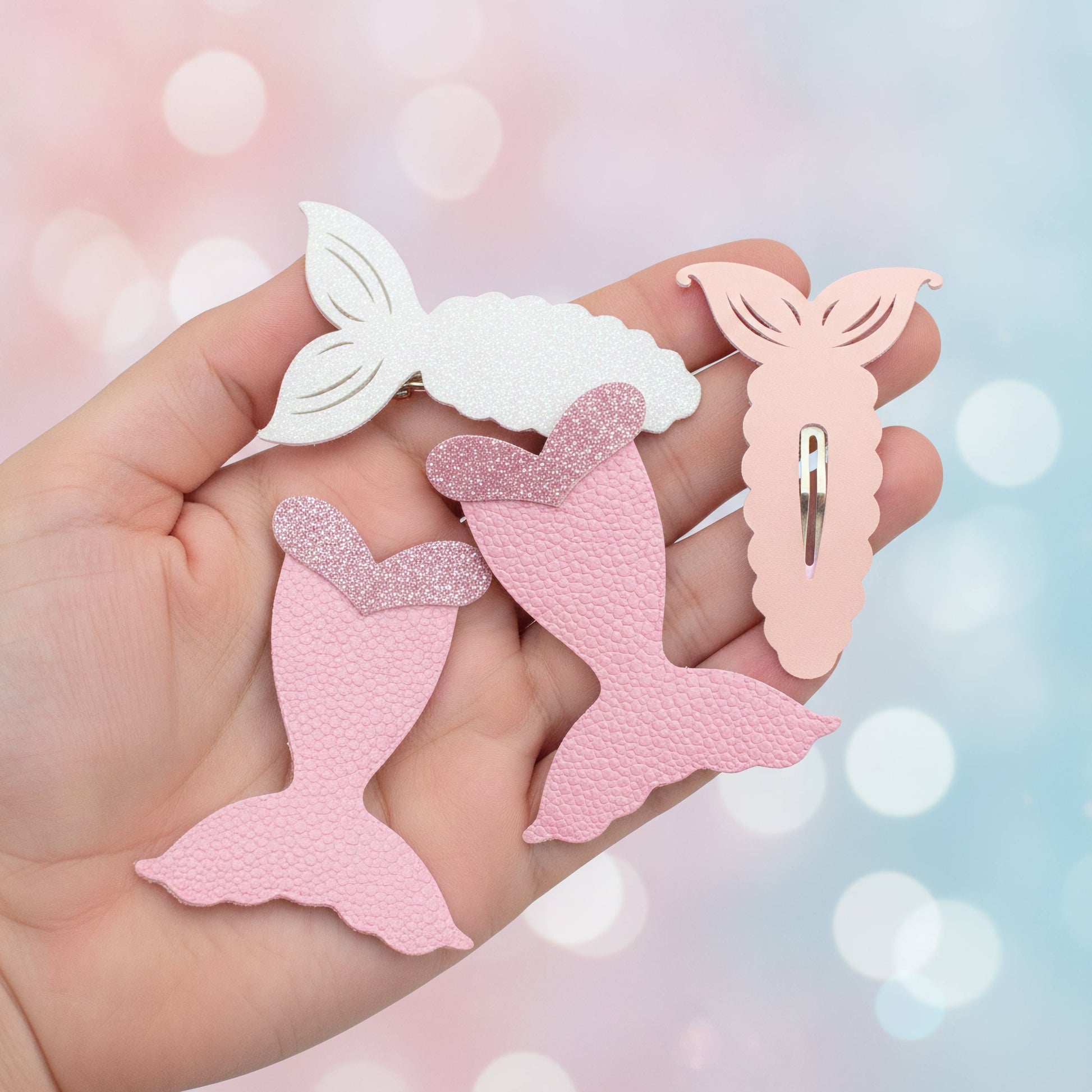 Three mermaid tail hair clips held in a hand against a soft pastel bokeh background. Two clips are pink, one is white, and the pink clips have glittery textured details.