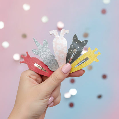 A hand with pink manicured nails holds four small hair claw clips shaped like animals and objects: a red snail, a silver rabbit, a white bunny with pink spots, a black cat, and a yellow giraffe. The background is a soft pink-to-blue gradient with blurred bokeh lights.
