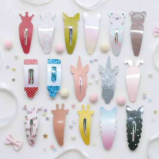 A flat-lay collection of twenty cute animal and unicorn-themed hair clips arranged on a white background with decorative ribbons, pearls, and star confetti. The clips feature various designs including gradients, glitter, floral prints, and animal silhouettes.