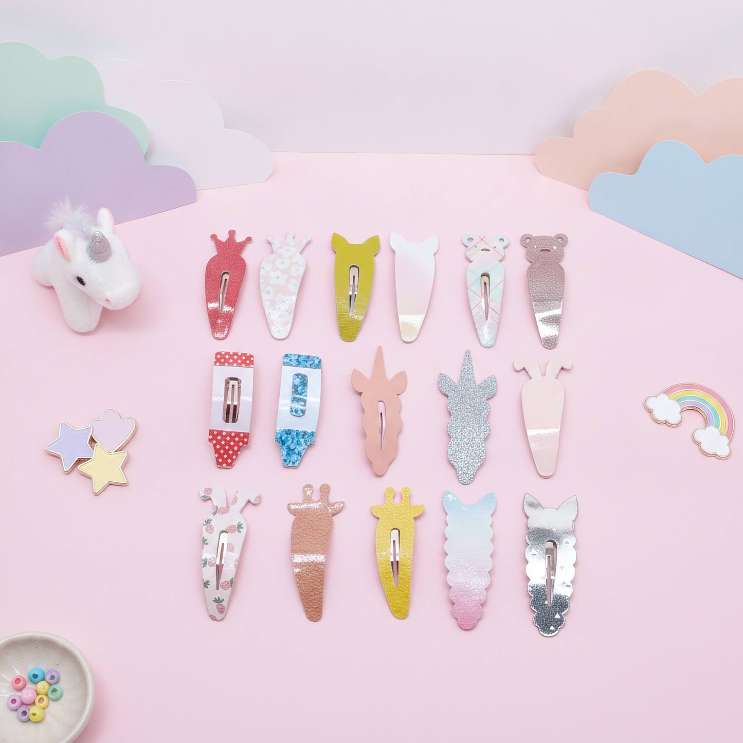 A set of pastel unicorn-themed hair clips arranged on a pink background with decorative cloud cutouts, a small unicorn figurine, stars, and a rainbow. The clips feature various pastel colors, glitter finishes, and cute unicorn designs.