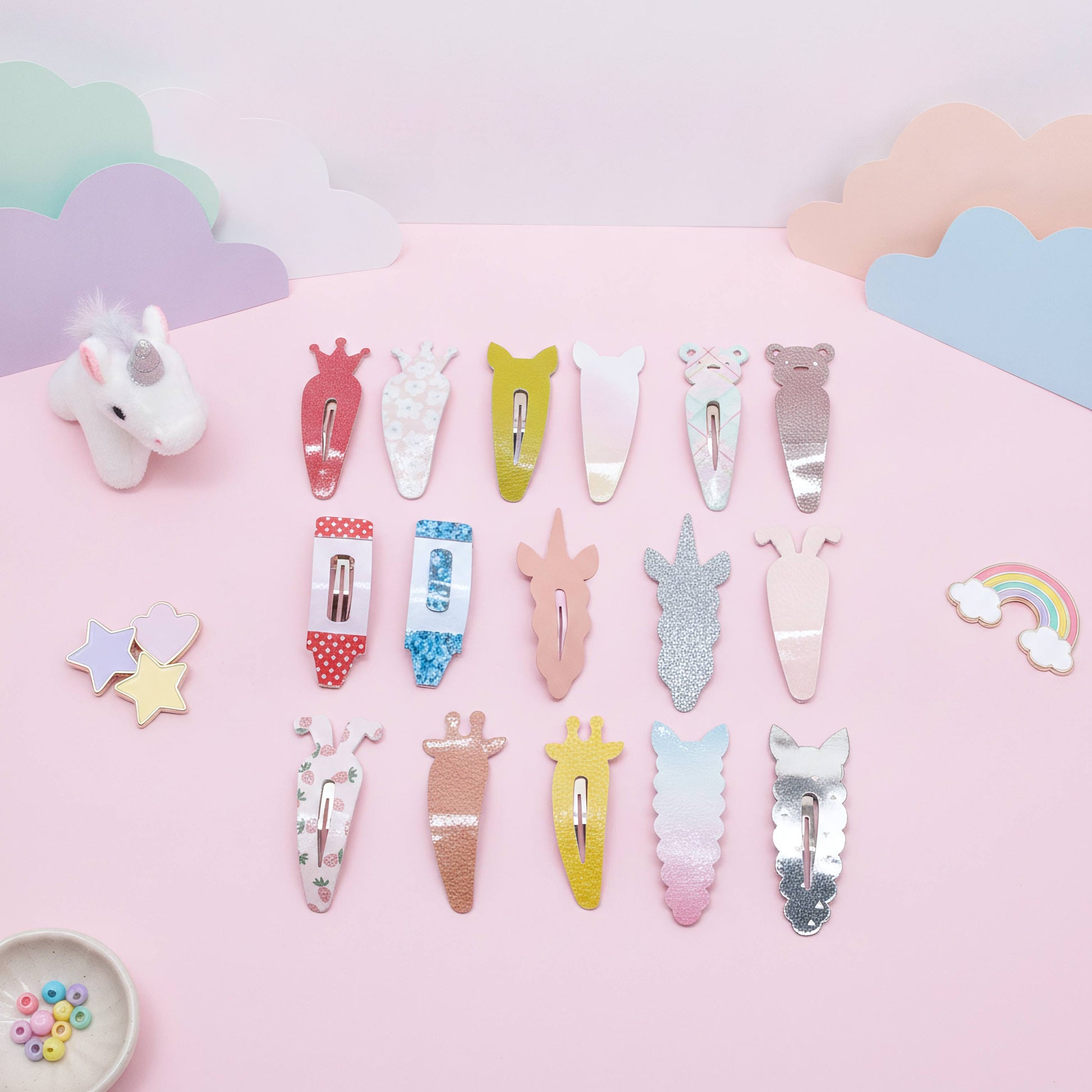 A set of pastel unicorn-themed hair clips arranged on a pink background with decorative cloud cutouts, a small unicorn figurine, stars, and a rainbow. The clips feature various pastel colors, glitter finishes, and cute unicorn designs.