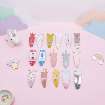 A set of pastel unicorn-themed hair clips arranged on a pink background with decorative cloud cutouts, a small unicorn figurine, stars, and a rainbow. The clips feature various pastel colors, glitter finishes, and cute unicorn designs.