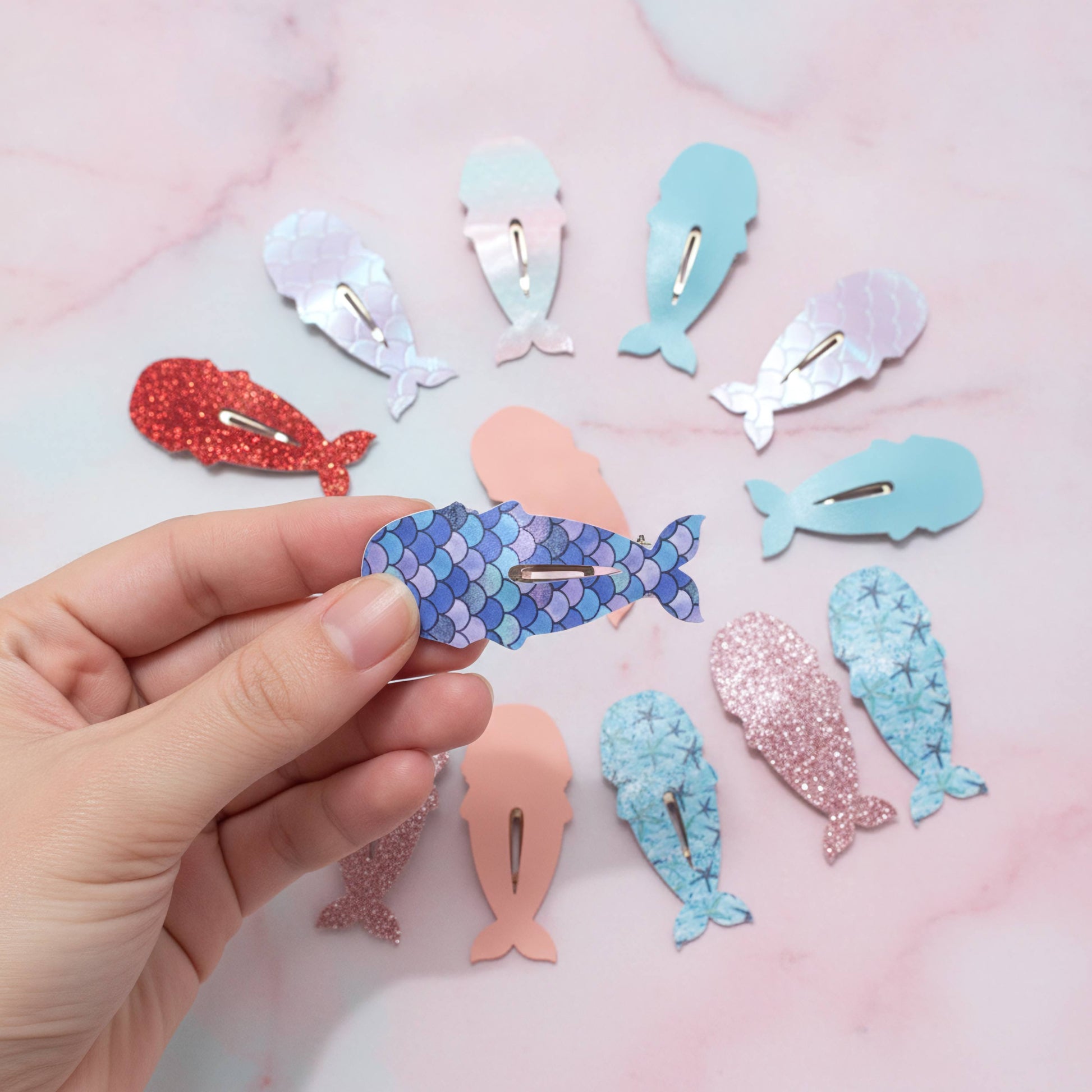 A hand holds a blue mermaid-scale whale hair clip against a pink marble background, with nine other whale-shaped hair clips in various colors and textures arranged around it.