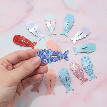 A hand holds a blue mermaid-scale whale hair clip against a pink marble background, with nine other whale-shaped hair clips in various colors and textures arranged around it.
