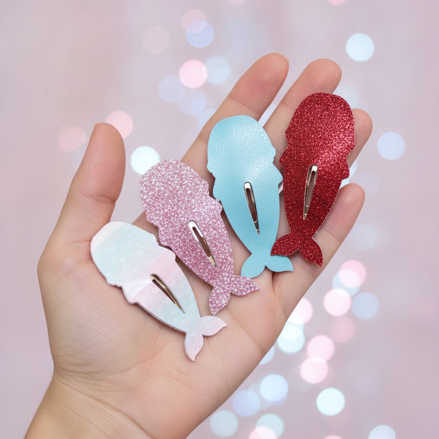 Four whale-shaped hair clips in pastel and glitter colors held in a hand against a soft pink bokeh background. The clips are white, pink glitter, light blue glitter, and red glitter.