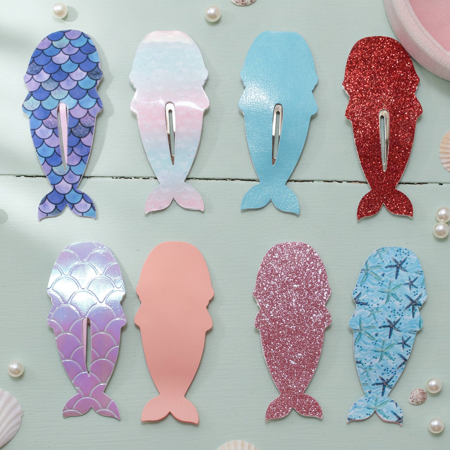 Eight mermaid-shaped hair claw clips arranged on a light blue wooden surface, each with a different pastel and glitter pattern including blue scales, pink and blue gradient, solid light blue, glittery red, iridescent purple, solid peach, glittery pink, and blue starfish print.
