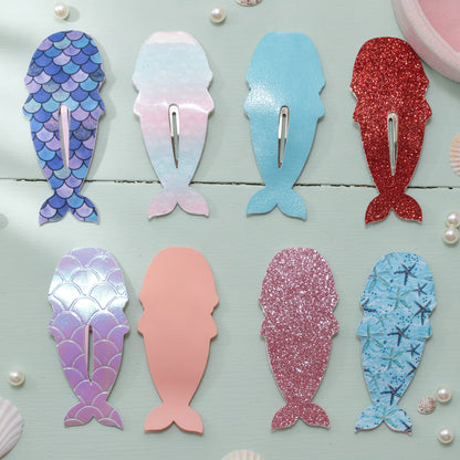 Eight mermaid-shaped hair claw clips arranged on a light blue wooden surface, each with a different pastel and glitter pattern including blue scales, pink and blue gradient, solid light blue, glittery red, iridescent purple, solid peach, glittery pink, and blue starfish print.