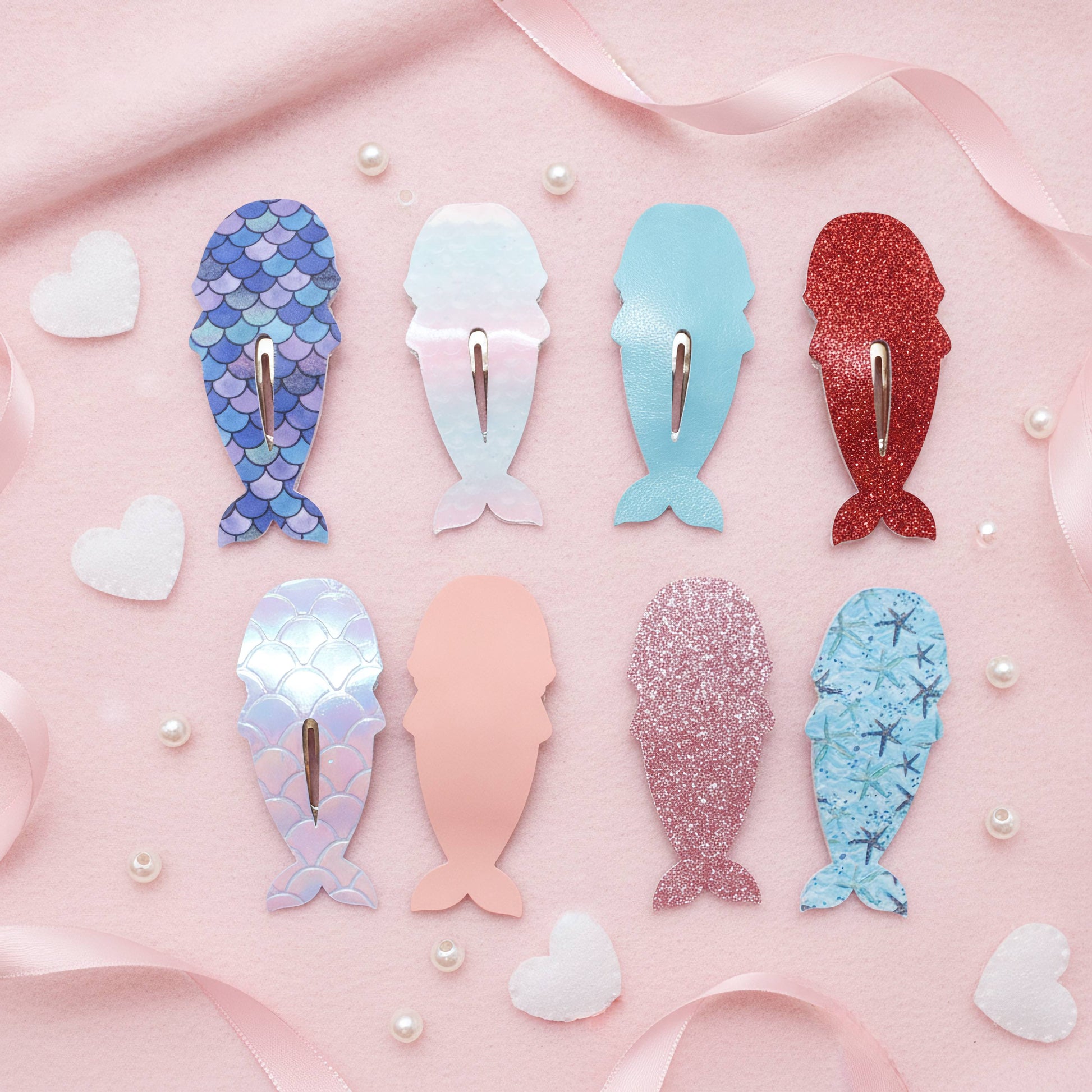 Eight mermaid-shaped hair clips arranged on a pink background with ribbon and heart decorations. The clips come in blue, pink, and red colors with various textures including scale pattern, glitter, holographic, and solid.
