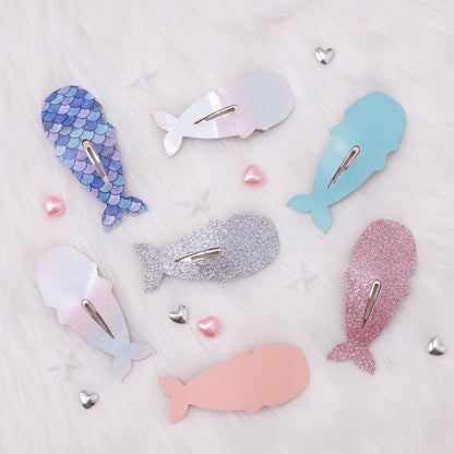 Six mermaid-themed whale hair clips in various colors and textures, including blue and purple scales, glitter, and solid pastel shades, arranged on a white faux fur background with small decorative hearts.