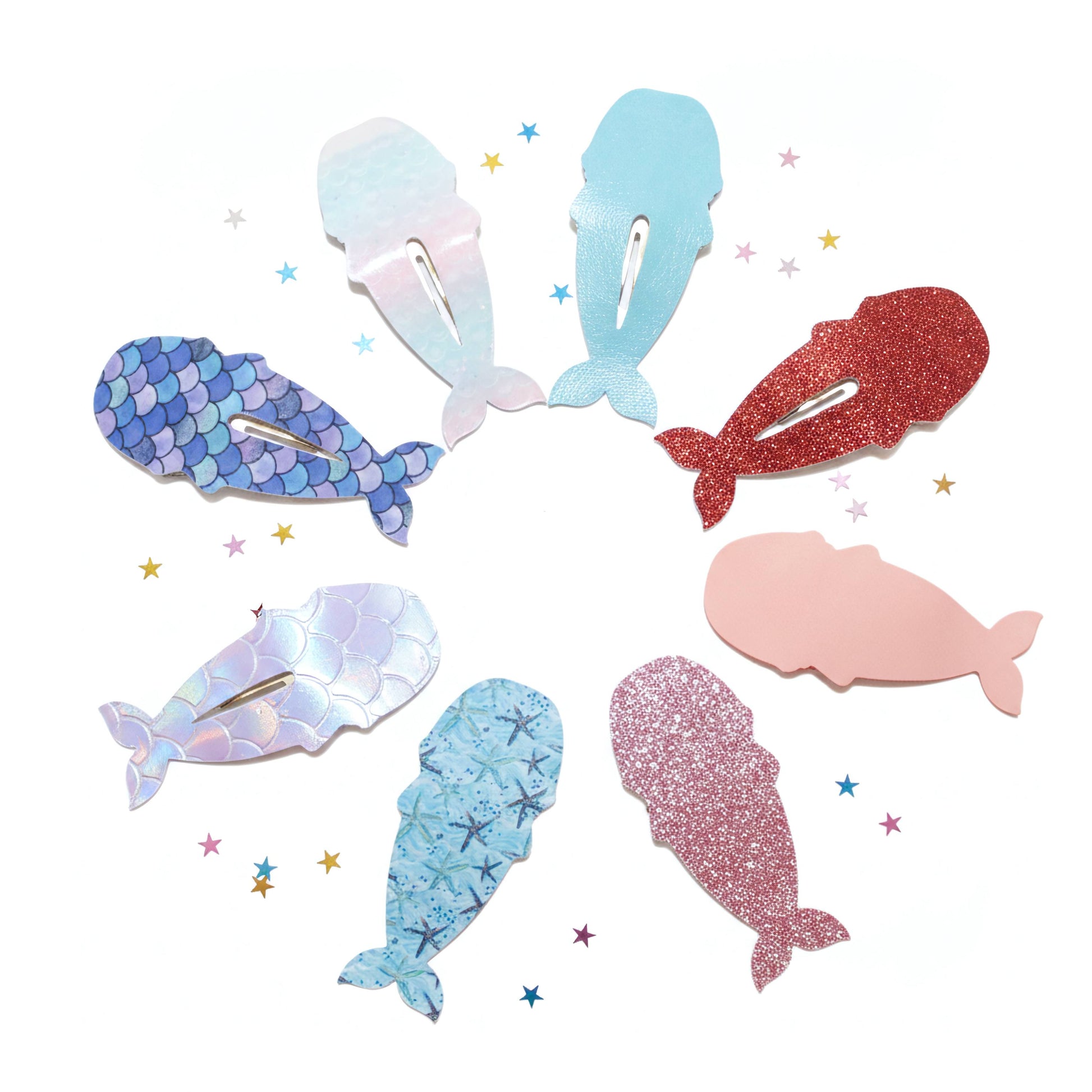 Seven whale-shaped hair clips in various colors and textures, including glitter, iridescent, and solid shades, arranged in a circle on a white background with small star accents.
