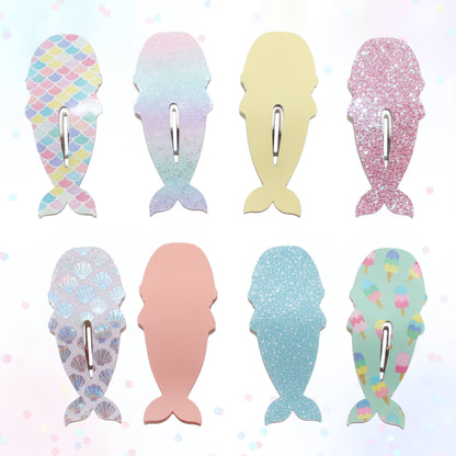 Eight mermaid tail-shaped hair clips arranged in a 2x4 grid on a light pastel background. The clips feature different designs: scales, iridescent gradient, solid pale yellow, glitter pink, scallop pattern, solid pale pink, glitter light blue, and ice cream print.