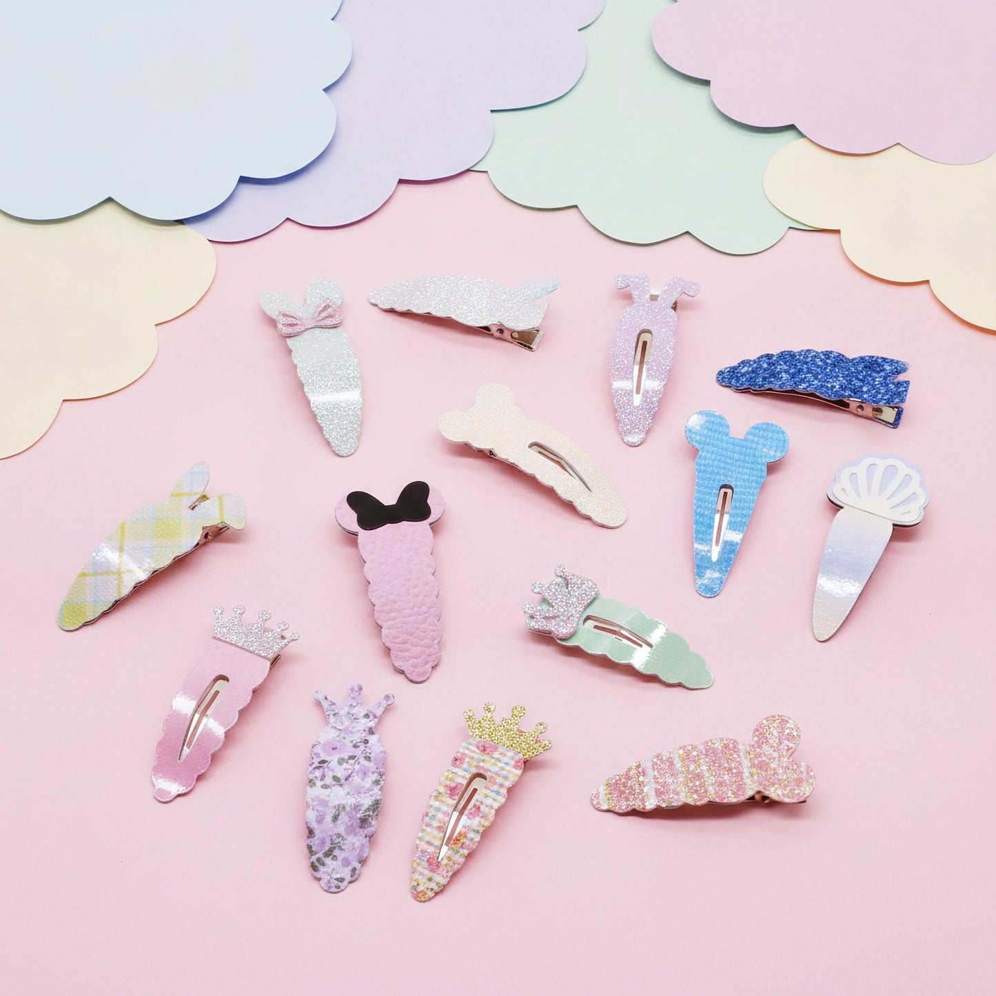 A set of pastel-colored hair clips arranged on a pink background with decorative cloud-shaped paper cutouts. The clips feature various designs including bows, flowers, shells, and glittery patterns in pink, blue, white, and yellow.